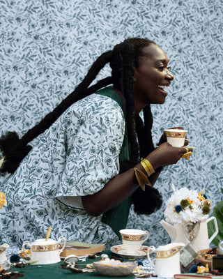 A happy dark skin woman in afrocentric hairstyle and jewellery drinking heritage tii from a bridgerton-inspired fancy tea set