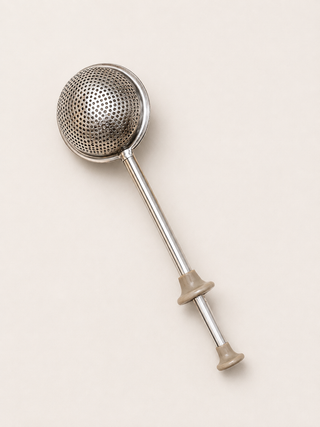 A stainless steel teabag infuser with a spherical mesh head and a long handle featuring beige plastic end caps, shown on a solid light beige background.