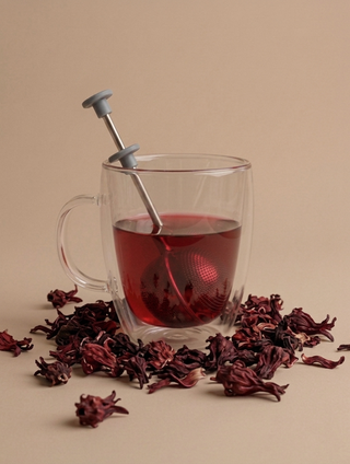 A clear glass mug filled with dark red tea and a metal tea infuser, surrounded by scattered dried red flowers on a beige surface, with dark brown letterbox bars at the top and bottom.